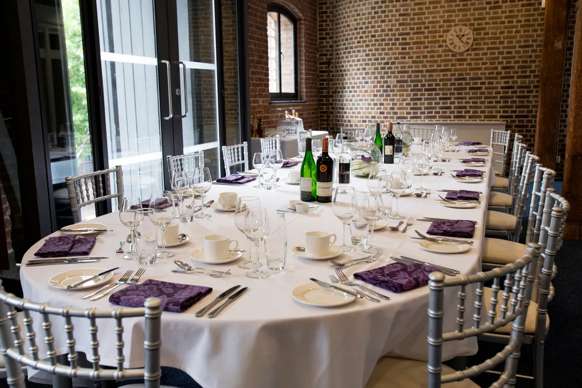 Elegant dining table set with white tablecloths, purple napkins, and an assortment of glassware and utensils, arranged for a formal meal, with bottled beverages and brick wall in the background.