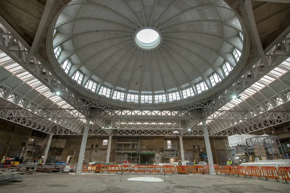 A large, empty market building under renovation with a circular dome ceiling, metal supports, and construction materials scattered around. Orange barriers cordon off parts of the site.