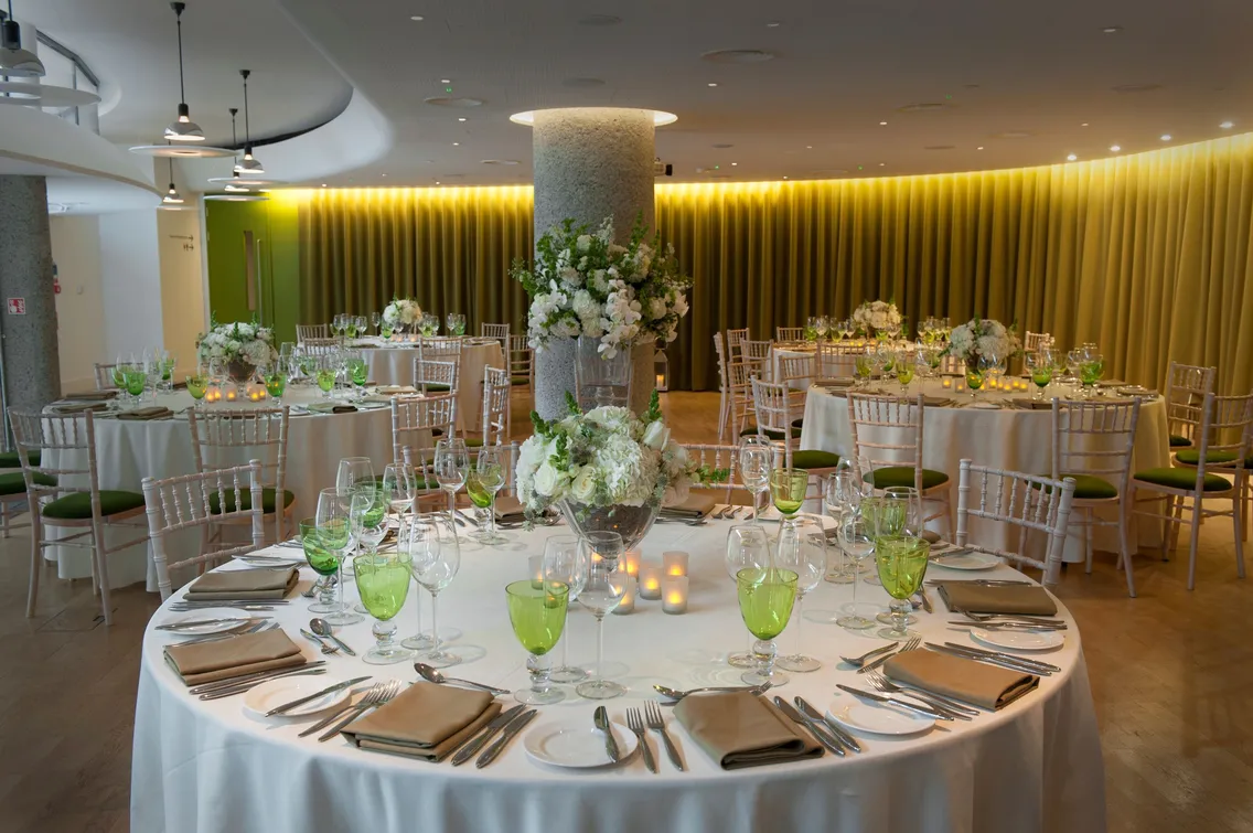 A well-lit banquet hall with circular tables set for an elegant event. Tables feature white linens, green glassware, flower centerpieces, and an arrangement of plates and silverware.